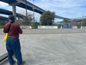 A man with his back to the camera, wearing a backward baseball cap, long-sleeved T-shirt and jeans looks over a concrete landscape under a highway, where slabs of graffitied cement line the far boundary.