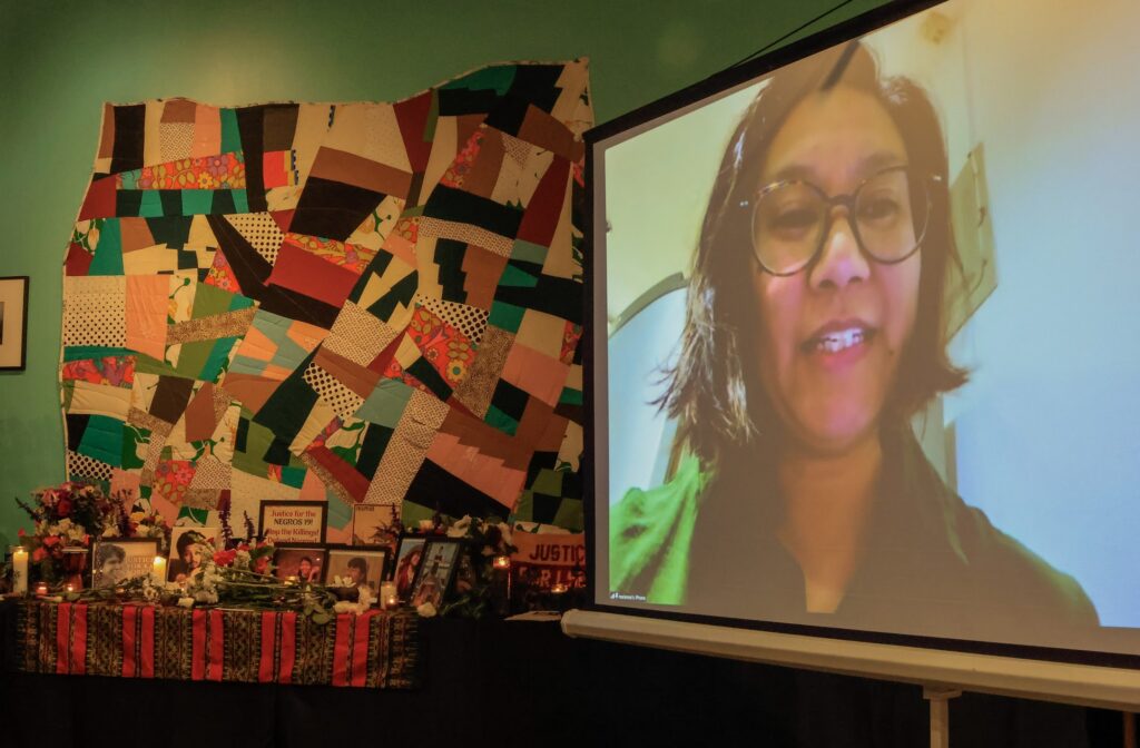 A woman's face is projected on a screen to the right of the frame. She has a short brown bob and is wearing large bone-framed glasses. To the left of the frame is a colorful tapestry tacked to a green wall. In front of it is a tribute table filled with photos and mementos.
