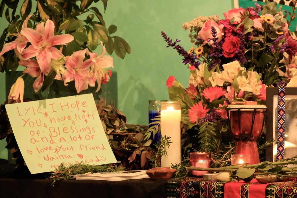 Lit candles of various sizes sit on an altar-like table in front of two vases of flowers. To the left is a hand-written sign in red crayon.