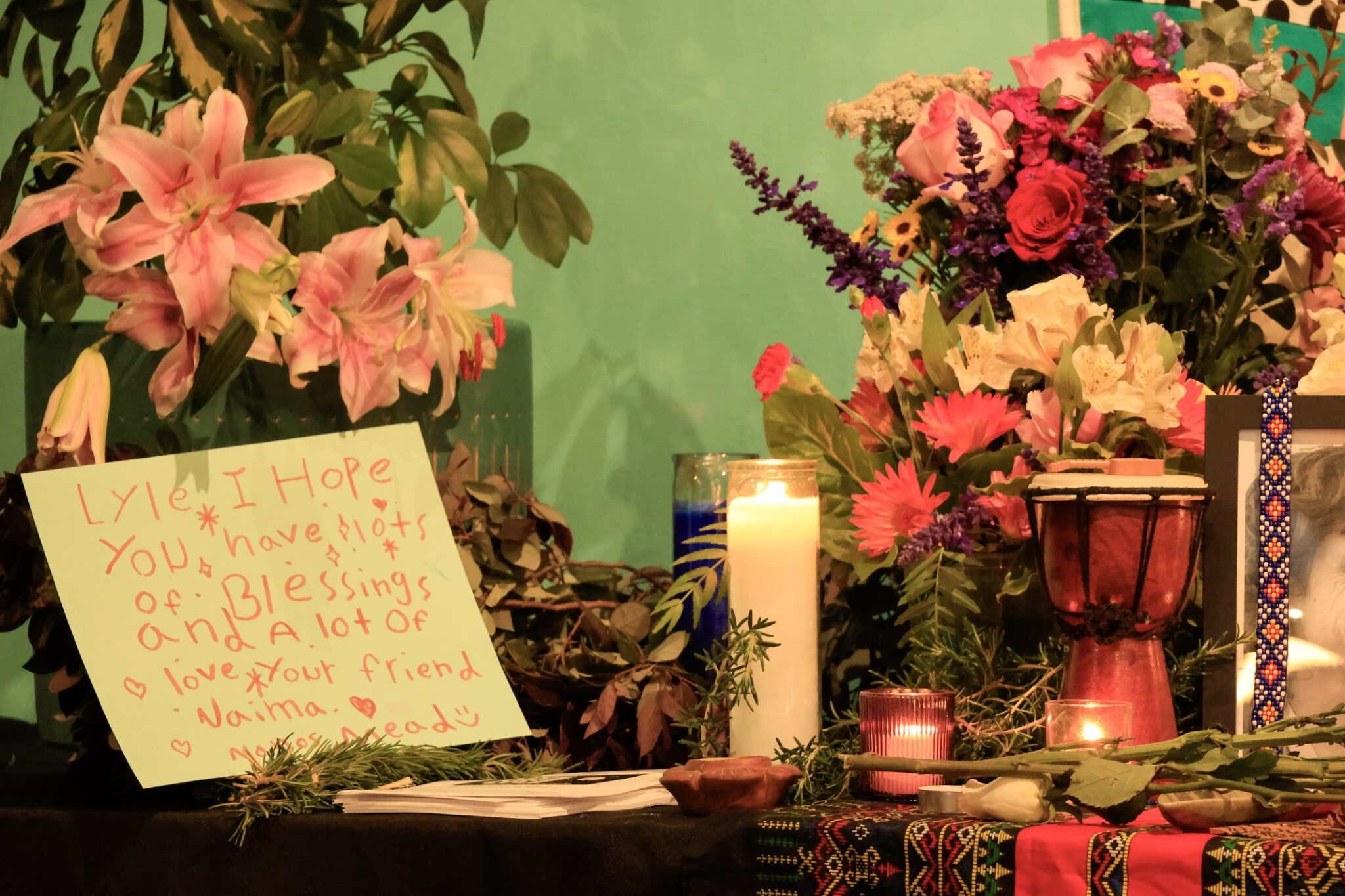Lit candles of various sizes sit on an altar-like table in front of two vases of flowers. To the left is a hand-written sign in red crayon.