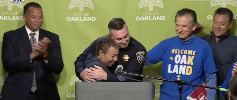 A tall police officer with cropped black hair hugs a small woman with short curly dark hair behind a podium and in front of a green City of Oakland banner, as two men stand to the right of the screen smiling and one man smiles on the left.