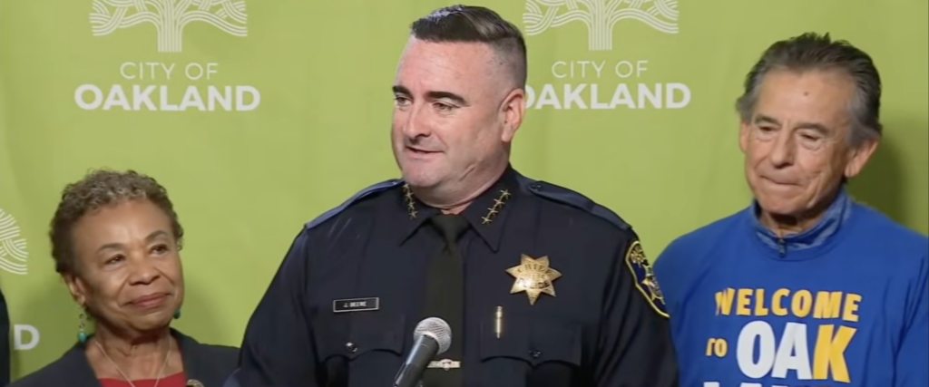 A police officer with cropped dark hair stands at a microphone between a short woman with short curly dark hair and an older man with short dark hair and a blue sweatshirt.