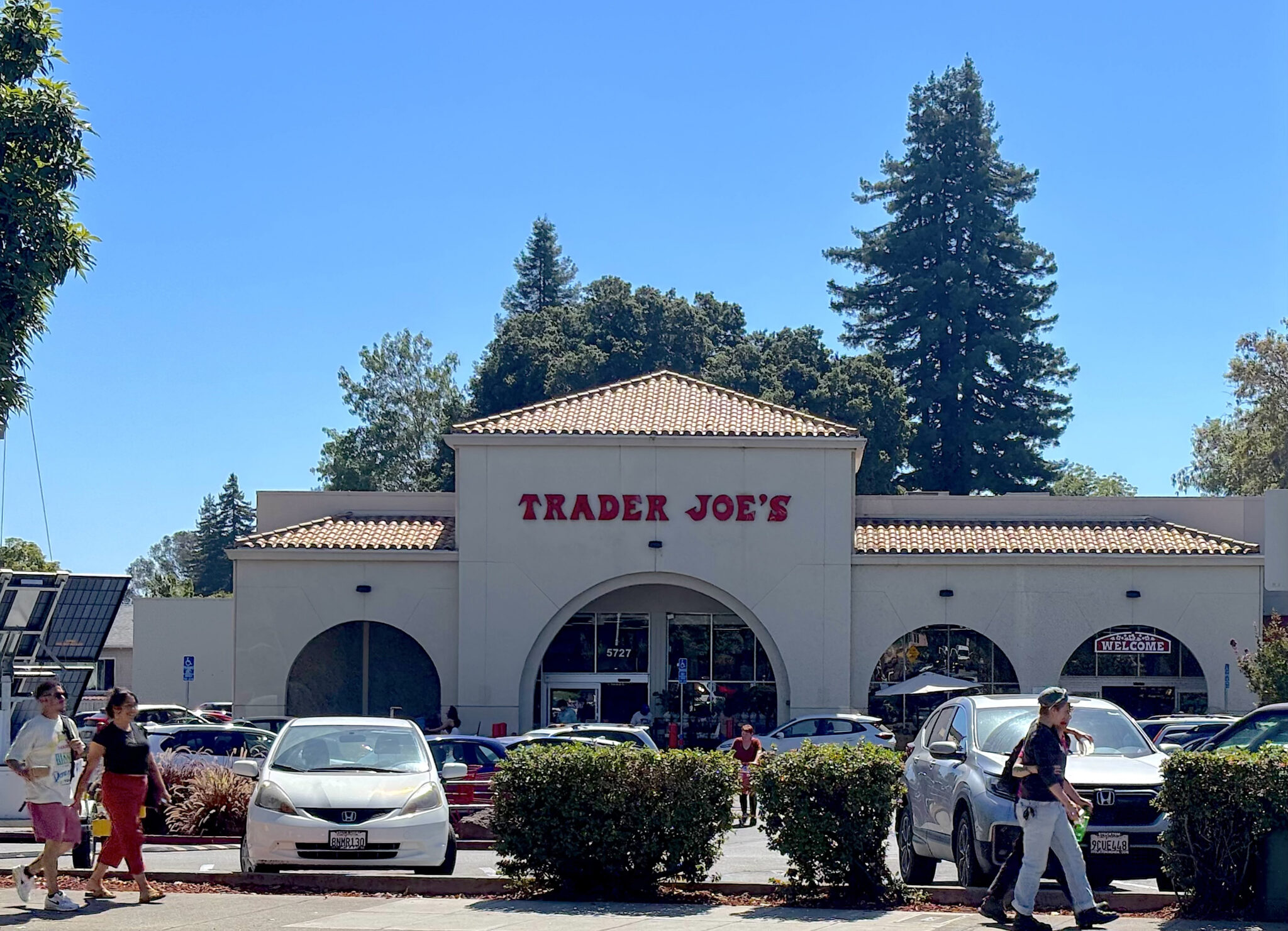 A one-story beige storefront with the store name in red letters sits behind several rows of cars in a parking lot and under a blue sky.