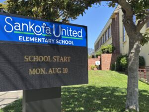 A closeup of a digital sign outside a school that says school start Mon. Aug. 10. It sits under a tree on grass next to a beige two-story building.