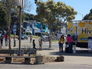 A wide shot of a corner of a cross road, in which a woman with a child is chatting with another woman by a yellow van