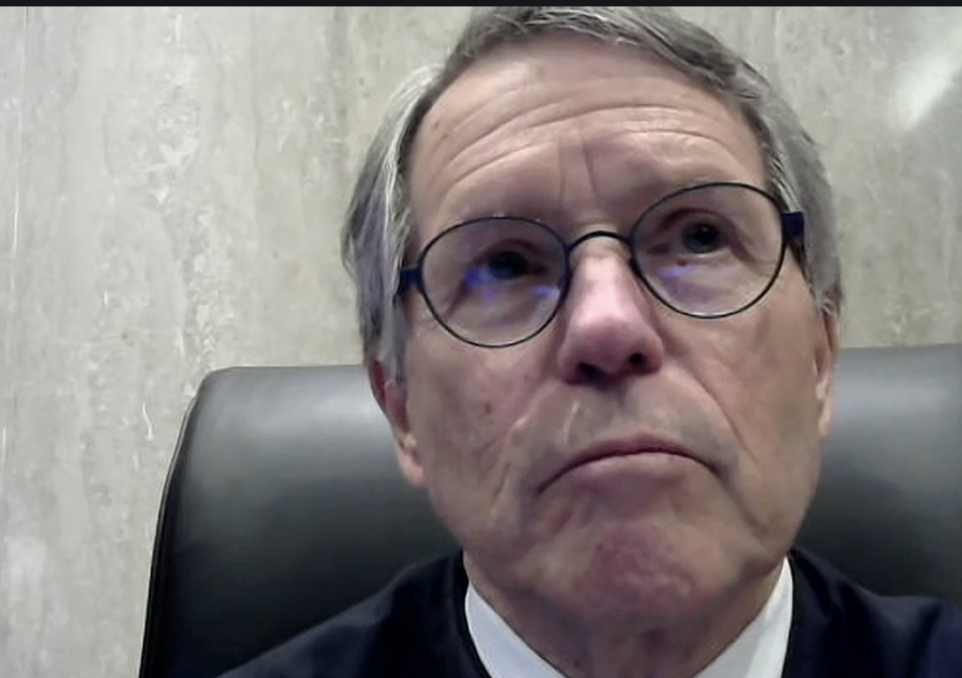 A closeup of a man's face. Short gray hair parted on the right side, black wire glasses, a couple moles on his cheek, sitting on a black leather chair, gazing toward the left.