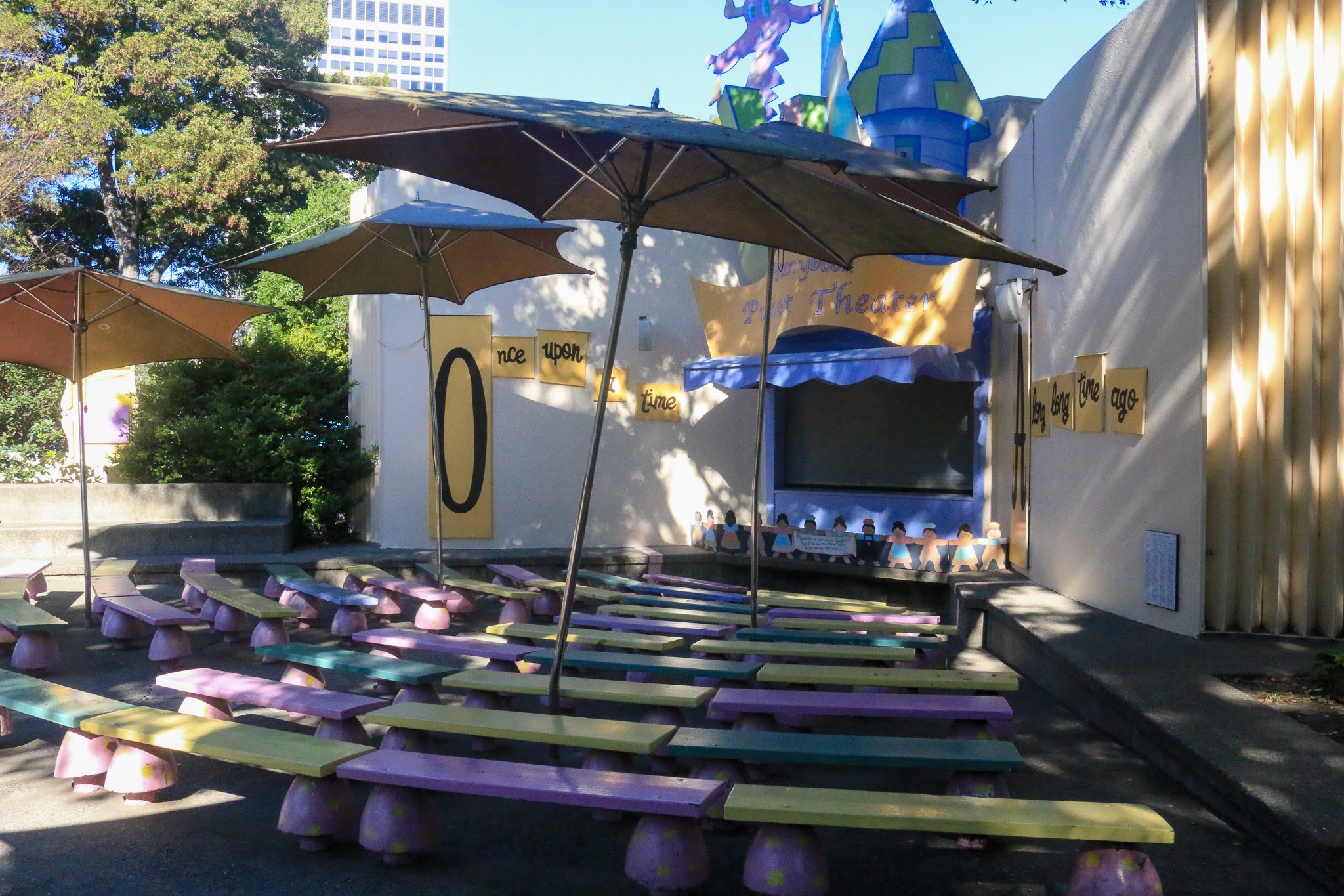 A small theater with about two dozen benches colores yellow, purple and green and a small blue stage for puppets. Three umbrellas shad the benches.