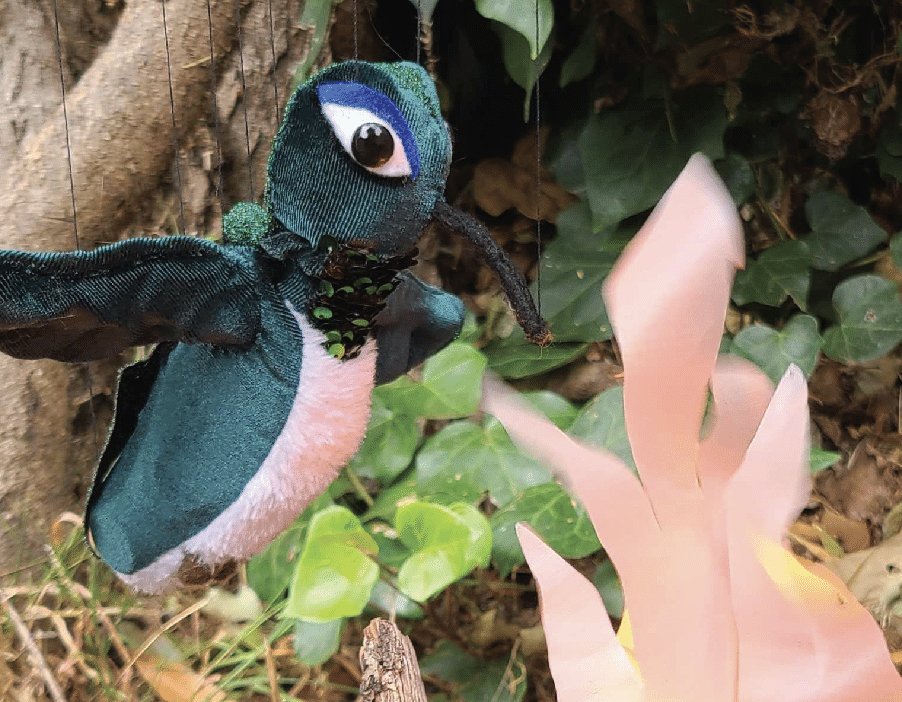 A blue hummingbird puppet made of cloth hovers by a pink flower and green leaves.