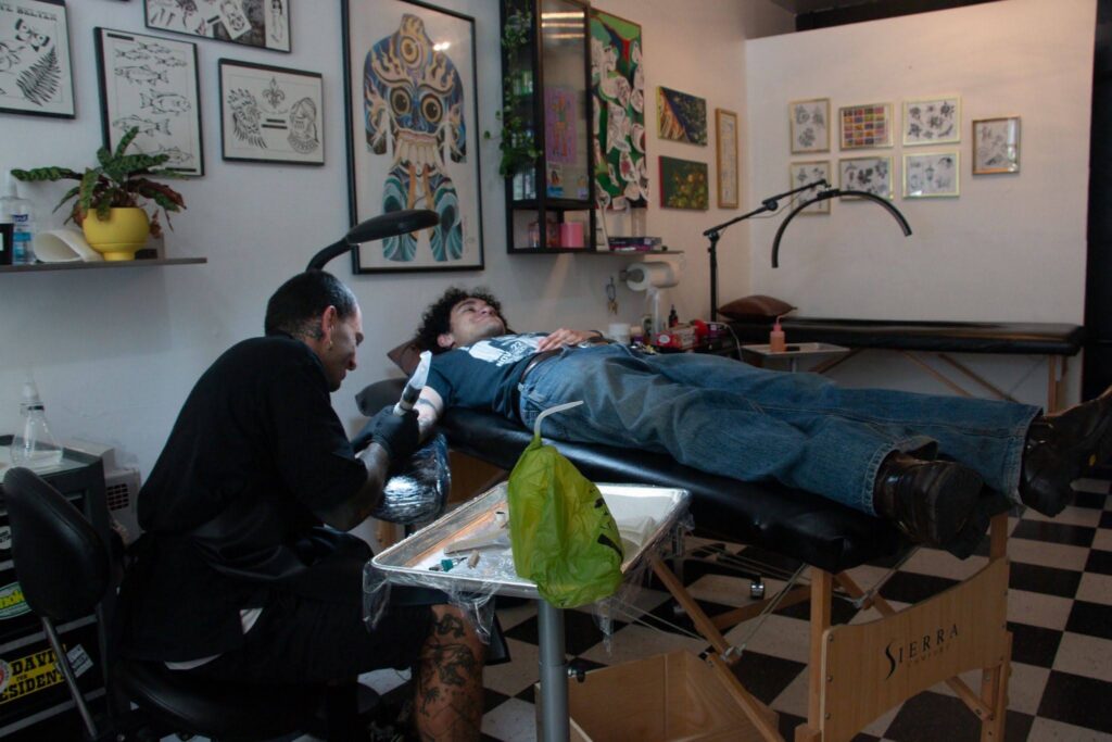 A wide shot of the tatoo shop, in which a middle-aged tattoo artist is tatooing on a young man with curly hair lying on a bed.