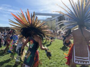 Two people, among a larger group in a circle, dressed with traditional Aztec costume, is dancing on the grass