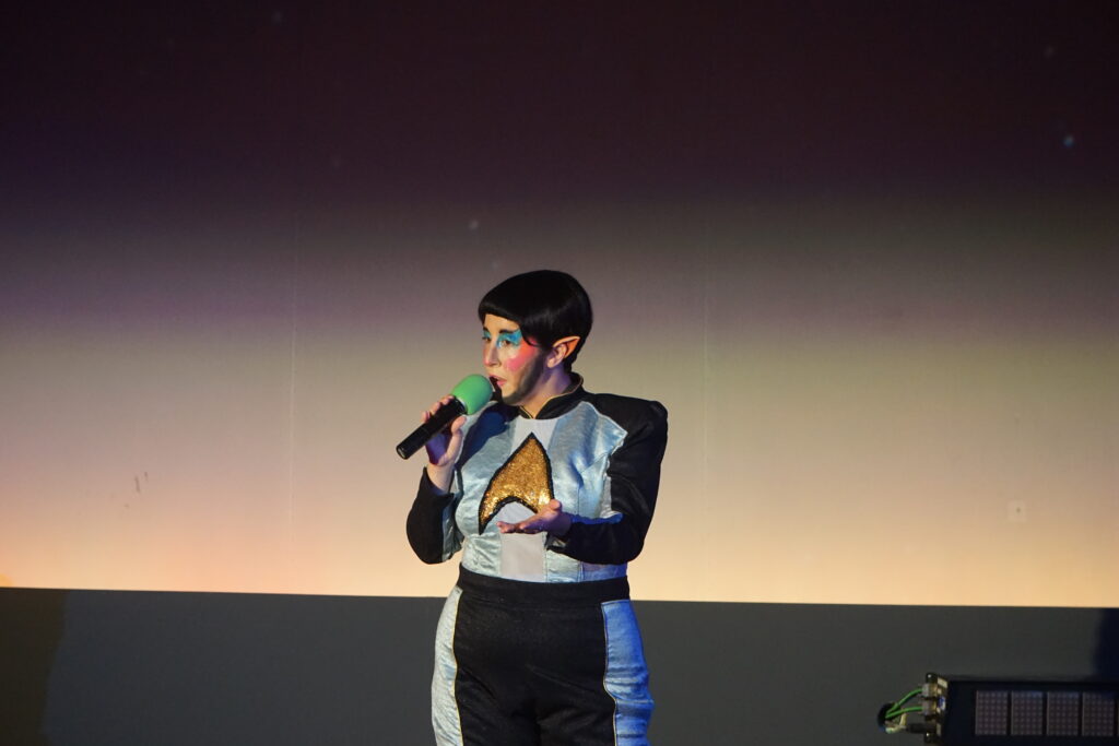 Drag king VERA! dressed as Mr. Spock, wearing pointy ears, colorful eye makeup, a black bowl-cut wig and a blue uniform featuring a gold emblem speaks into a microphone on stage at Chabot Space and Science Center.