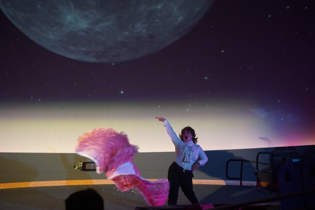 Jota Mercury on stage tosses a large pink feathered accessory into the air while singing. Behind them, a large screen projects an image of partial moon against a starry night sky.
