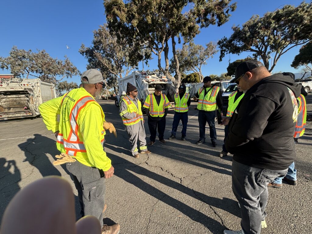 Eight cleaning workers gather in a circle and talking to each other in a parking lot. Behind them are three trees and a garbage truck.