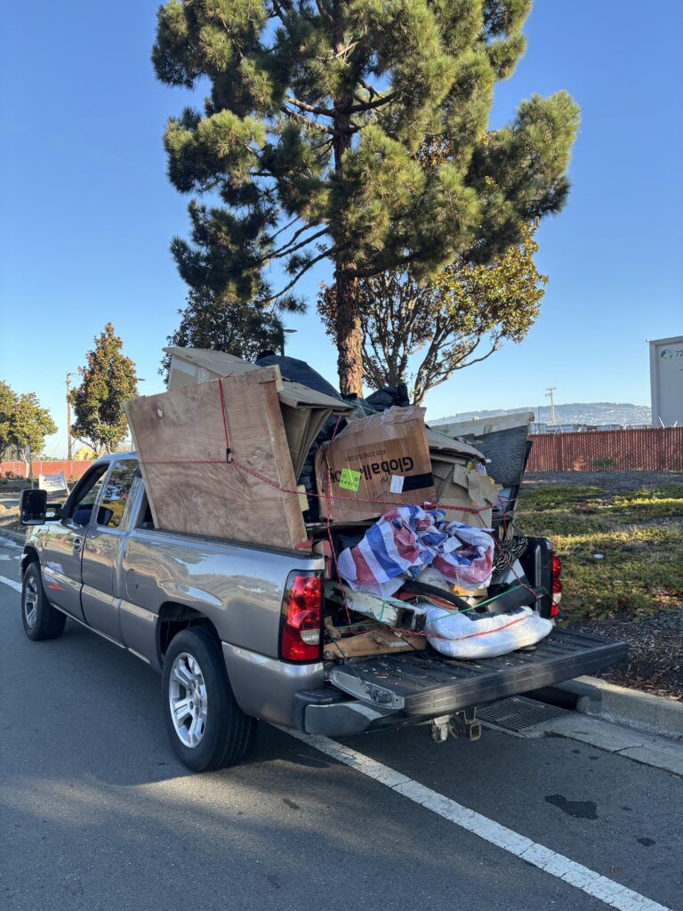 A silver pickup truck loaded with bulky wastes pulled over by the curb, next to a pine tree.