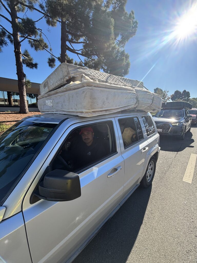 A man drives silver SUV waiting at the front of a line. Two damaged mattress is tied on top of the SUV.