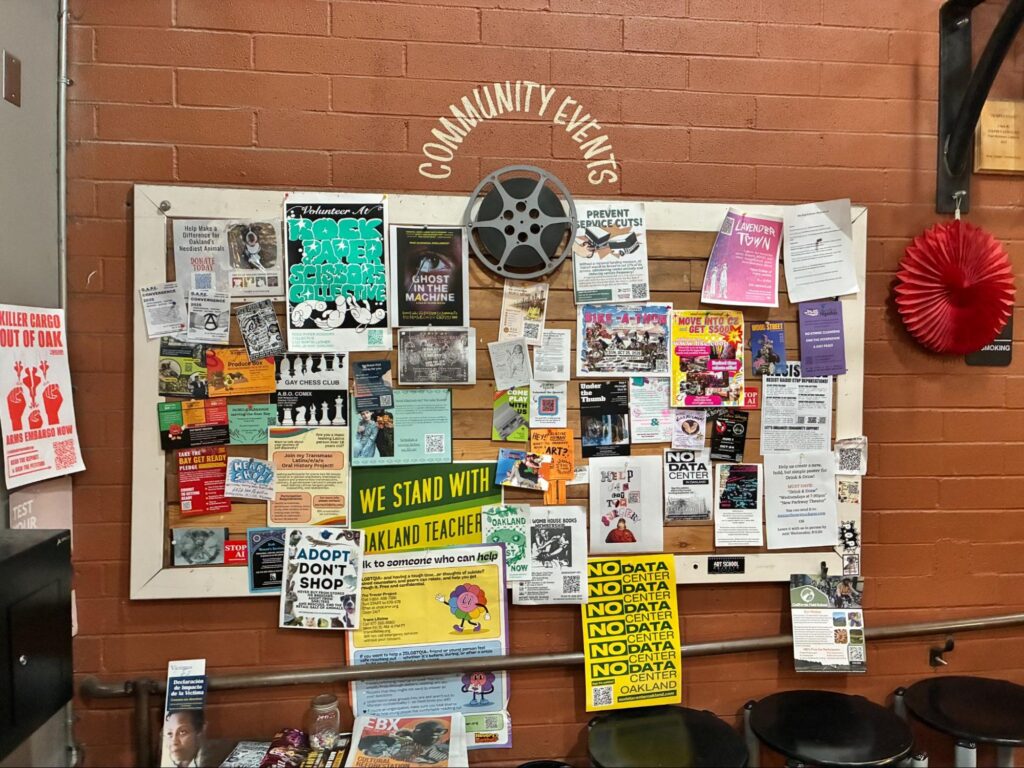 A framed community bulletin board mounted on an exposed red brick wall with the words "Community Events." The board is densely covered in pinned flyers, activist posters, and notices, including ones reading "We Stand With Oakland Teachers," "Adopt Don't Shop," and "No Data Center".