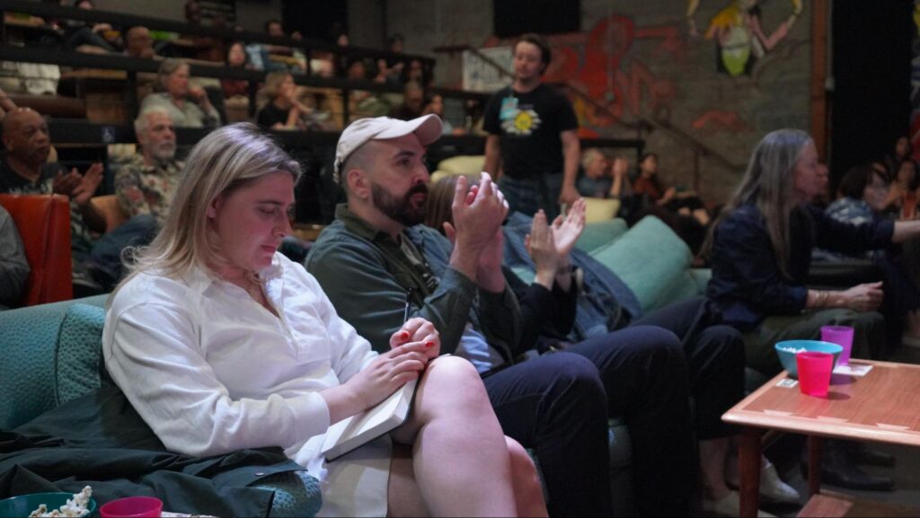A close-up view of attendees in an auditorium. In the foreground, a blonde woman in a white shirt and dark red nail paint takes notes with a pen in a notepad while seated on a teal couch. Next to her, a man in a cap and other audience members applaud towards the stage as a man wearing a badge walks by in the background.