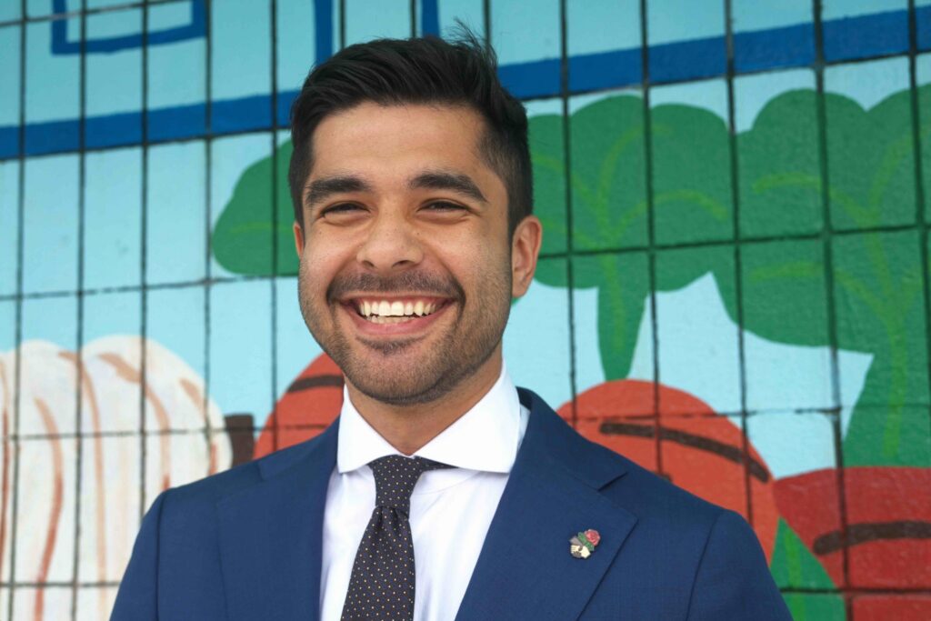 Outdoor portrait of Oakland District 2 candidate Nate Adams smiling warmly in a navy suit and patterned tie against a colorful tiled wall mural.