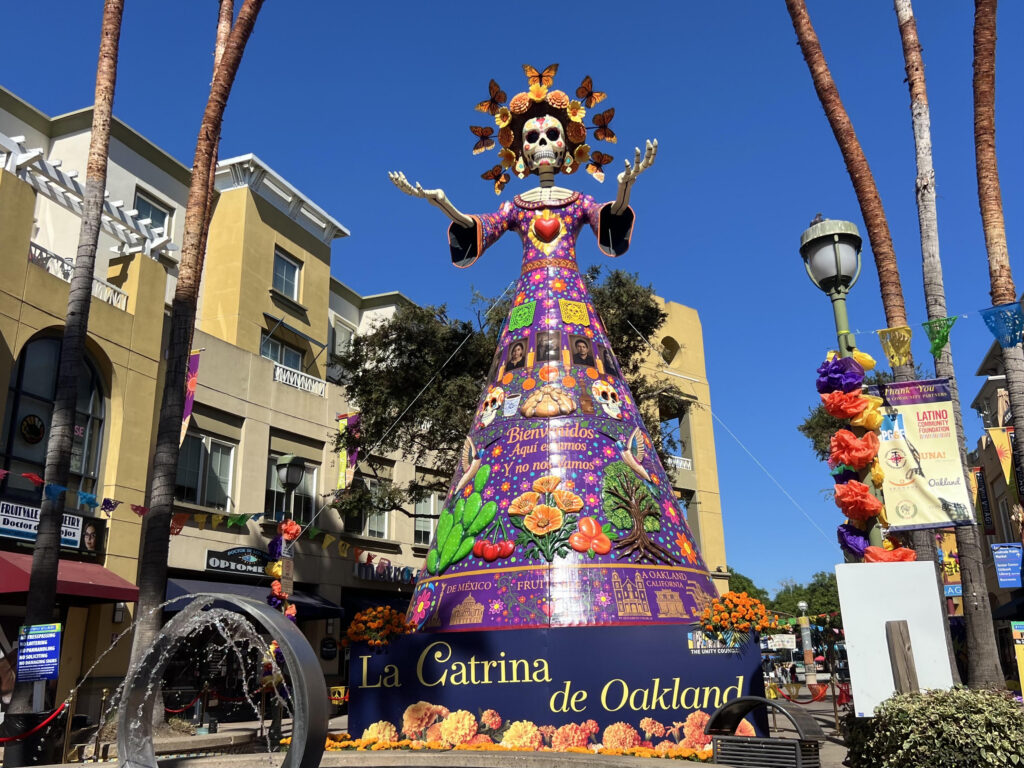 A colossal colorful female skeleton wearing an ornate hat stands on a blue base on a city street under a blue sky.