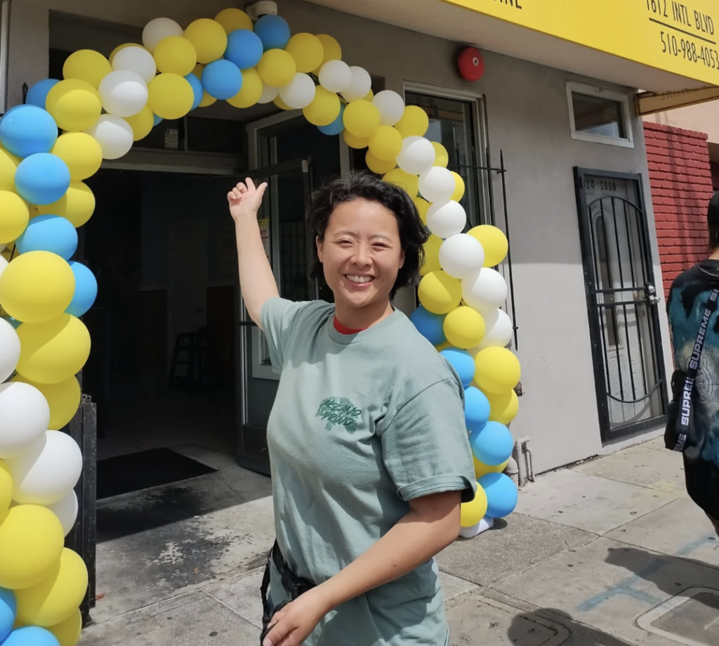 Oakland District 2 candidate Charlene Wang smiling and gesturing towards a yellow, blue, and white balloon arch framing a building entrance