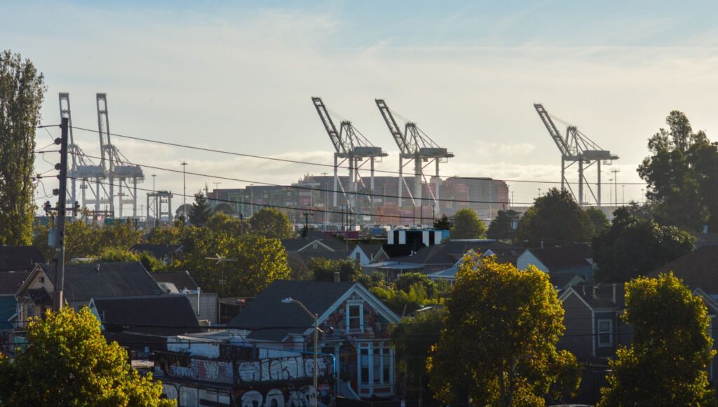 Port of Oakland with shipping containers, cranes and and a cargo ship under a pale sky with a residential West Oakland neighborhood with homes and trees in the foreground.