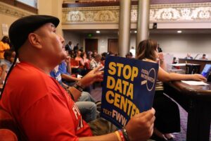 Seated in the audience section of Oakland City Council Chambers, a man wearing a black flat cap and red shirt holds up a blue poster sign reading "STOP DATA CENTER" with the Food & Water Watch logo visible.