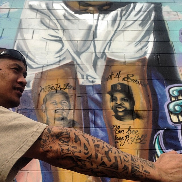 Close-up of a man wearing sunglasses on his head extending his forearm toward the camera to display a detailed black-and-grey tattoo spelling "DREAM" in graffiti lettering. In the background, a painted brick mural of a person displays portraits of two men on their hand.