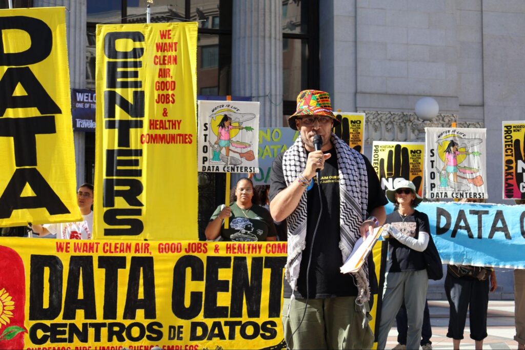Outdoors in front of Oakland City Hall, a man wearing a patterned bucket hat and a keffiyeh holds a clipboard and speaks into a microphone. Behind him, demonstrators hold large yellow and blue protest banners with slogans such as "DATA CENTERS" and "WE WANT CLEAN AIR, GOOD JOBS & HEALTHY COMMUNITIES."