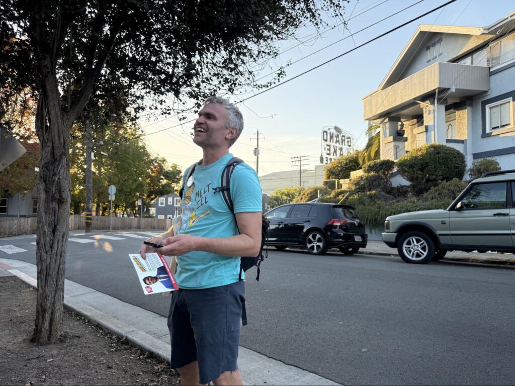 A man with short graying hair wearing a light blue "MELT ICE" t-shirt, dark shorts and a backpack stands on a sidewalk corner holding campaign flyers of District 2 candidate Nate Adams. He smiles while looking up toward the street, with the Grand Lake Theatre sign visible in the background.