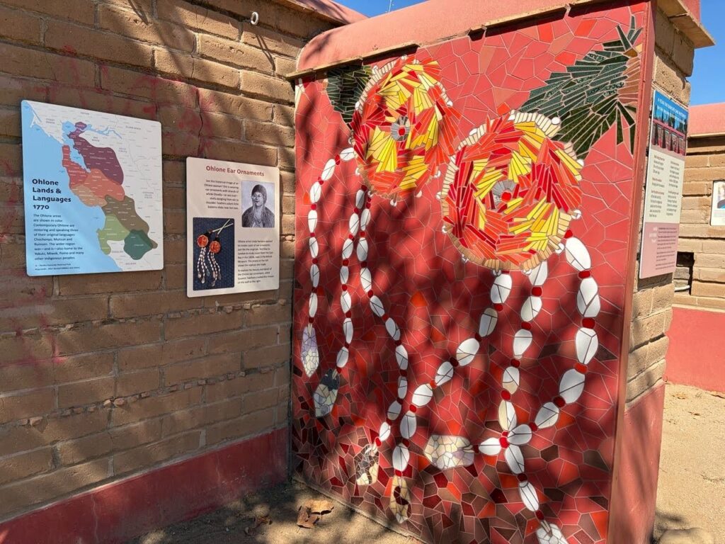 White shell-like pieces and red and yellow tiles make swervy lines in a mosaic on the side of a narrow brick wall.