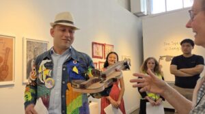 In a gallery a man with beige hat and colorful shirt holds a woodburned violin and shows it to a woman in glasses