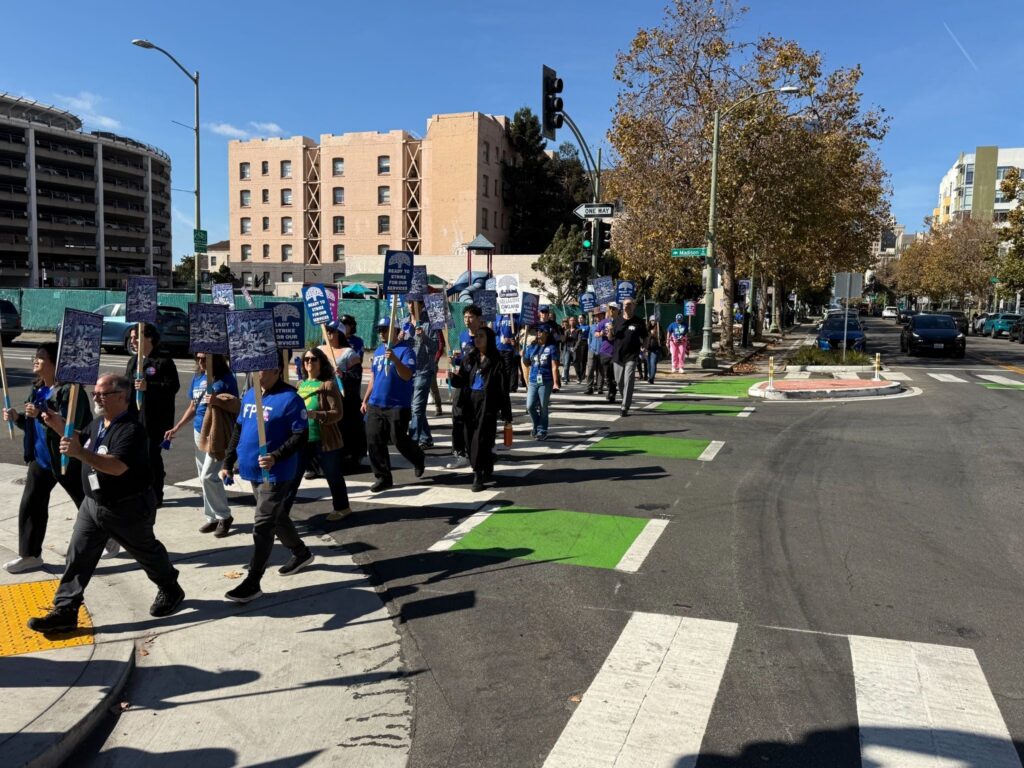 A few dozen people in blue T-shirts holding signs walk across a city street while a traffic light flashes green.