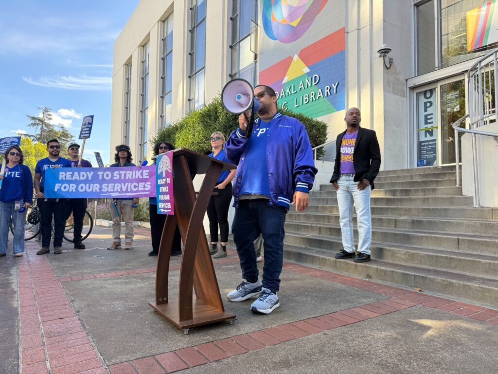 A man in a blue jacket speaks into a megaphone while a few people beside him stand with a banner. Behind him are steps leading to a white slabbed buiding.