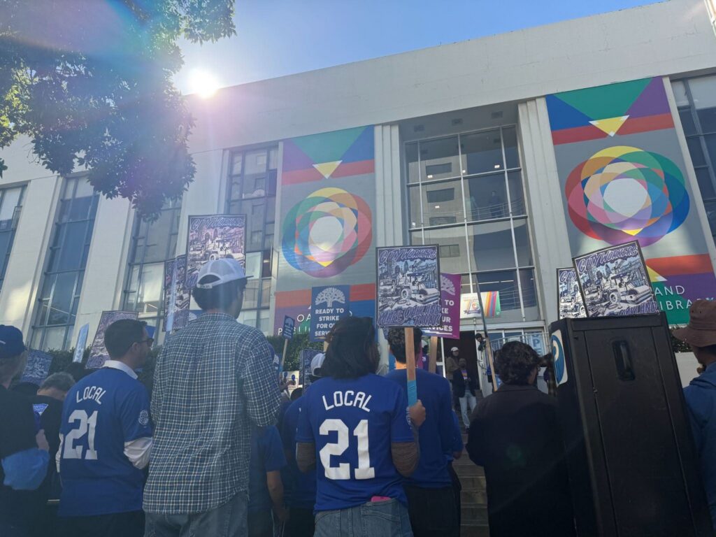 In front of a buidling with colorful panels, a group stands with signs, many wearing blue matching T-shirts with their union's name on the back.